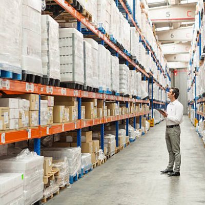 Full length of supervisor examining stock arranged on shelves in warehouse
